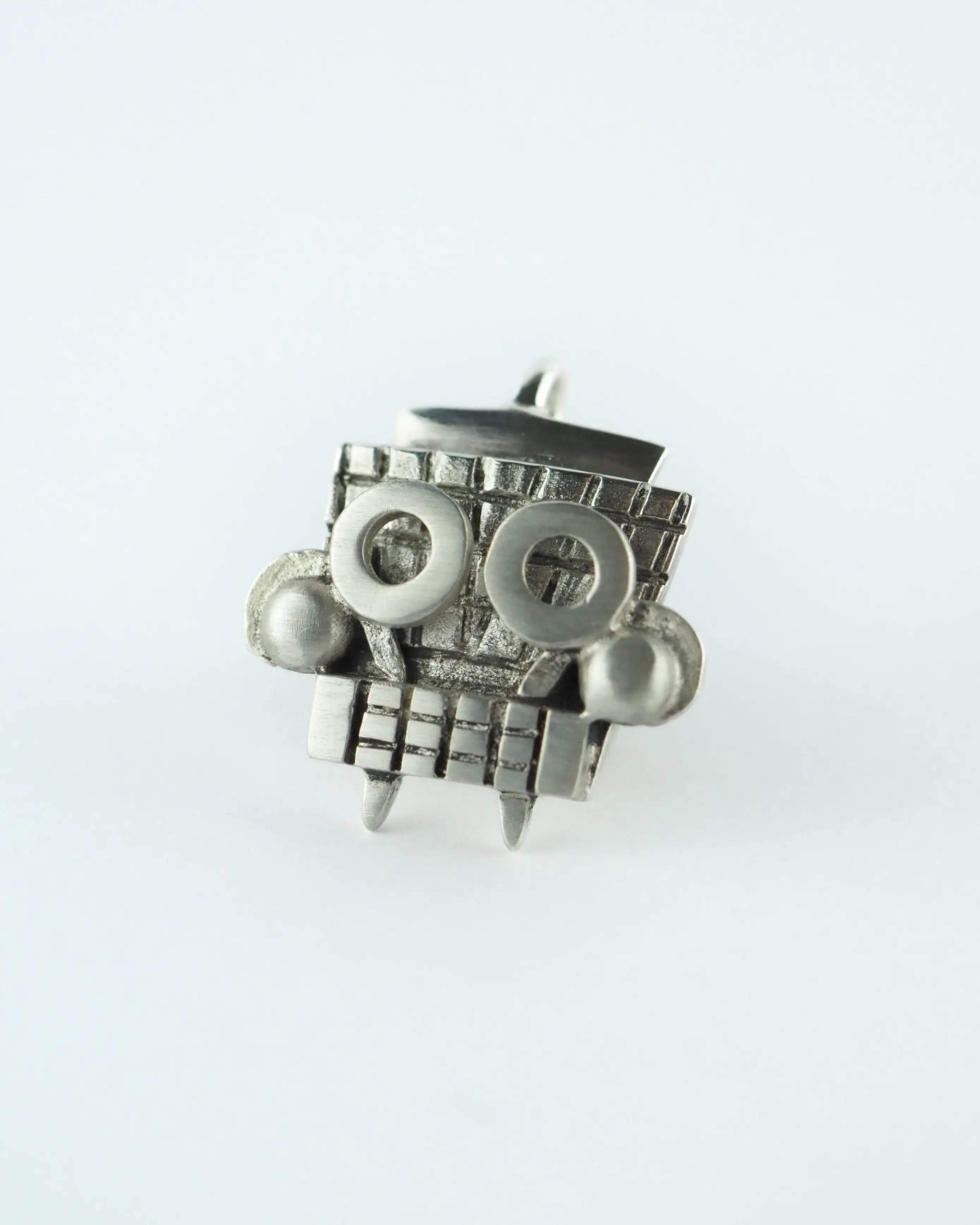 Colgante plateado de metal con figura tipo tótem y detalles enrejados